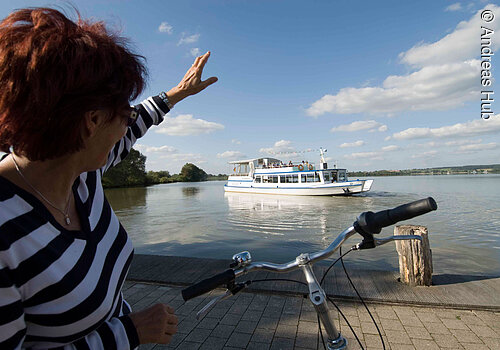 Eine Fahrradfahrerin winkt vom Ufer aus der MS Altmühlsee zu.