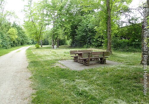 Unter dem Bäumen steht eine gemütliche Sitzgelegenheit aus Holz. Links führt ein geschotterter Weg entlang.