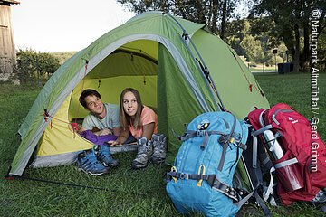 Eine Frau und ein Mann liegen im Zelt und schauen aus diesem heraus. Vor dem Zelt liegen ihre zwei Wanderrücksäcke.