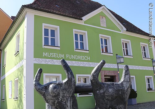 Außenansicht des Archäologischen Museums in Thalmässing. Grünes Haus mit schwarzem Dach. Davor steht eine Skulptur von menschlichen Figuren, die eine Platte tragen.