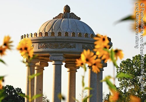 Fokus auf einen runden Steinpavillion im Schlosspark Dennenlohe. im Vordergrund gelbe Blumen.