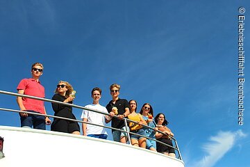 Eine Gruppe von sieben Jugendlichen lehnt an einer Schiffsreling und blickt nach schräg unten in die Kamera. Die Jugendlichen tragen Sonnenbrillen, Sommerkleidung und in ihrer Hand ein Getränk.