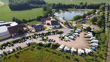 Luftaufnahme der Weißbachmühle. Von Links nach rechts erstreckt sich ein Pkw Parkplatz, dann folgen im Halbkreis angeordnetet Wohnmobile. Hinter dem Parkplatz stehen drei große Häuser in einer Reihe. Im Hintergund liegt das Naturfreibad umgeben von grünen Bäumen und Wiesen.