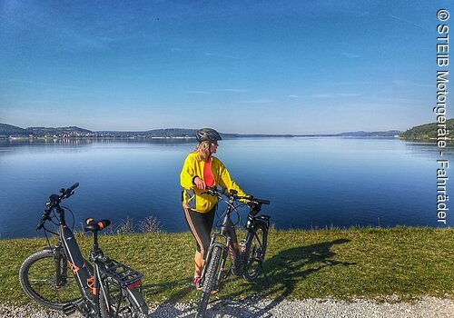 Eine Frau steht neben ihrem Fahrrad am Ufer eines Sees. Neben ihr steht ein weiteres Fahrrad. Hinter ihr ein blauer See unter blauem Himmel.