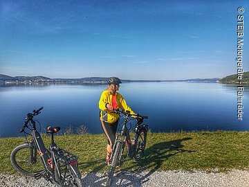 Eine Frau steht neben ihrem Fahrrad am Ufer eines Sees. Neben ihr steht ein weiteres Fahrrad. Hinter ihr ein blauer See unter blauem Himmel.
