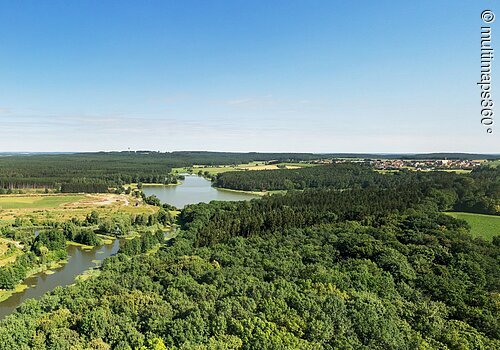 Panoramaaufnahme vom Denneloher See. Der See ist umgeben von grünen Wiesen und Wäldern.
