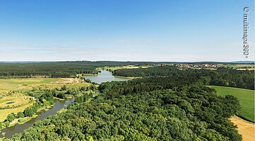 Panoramaaufnahme vom Denneloher See. Der See ist umgeben von grünen Wiesen und Wäldern.