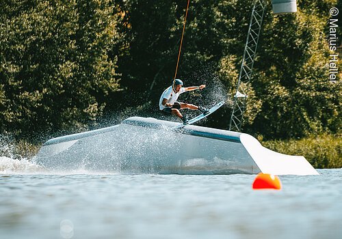 Ein Wakeboarder fährt über ein Hindernis im Wasser. Dahinter am Ufer grüne Bäume und Sträucher.