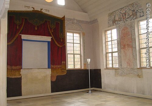 Innenraum einer Synagoge. Weiße Wände. Darauf ein roter gemalter Vorhang.