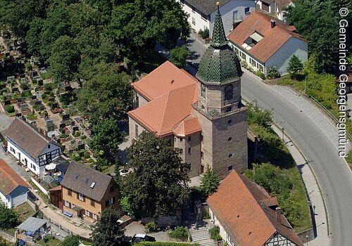 Luftbild eines Dorfes. In der Mitte eine Kirche mit grünem Turmdach. Drum herum Häuser und Bäume.
