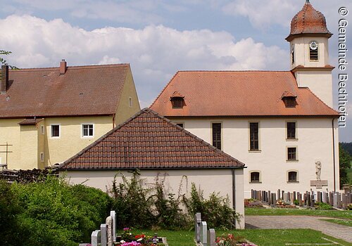 Eine Kirche mit Friedhof. Eine Kirche mit heller Fassade und kelinem Türmchen. Davor ein Friedhof mit Grabsteinen.