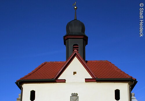 Ein Torbogen mit weißer Fassade und rotem Ziegeldach. Auf dem Dach ein kleines schwarzes Türmchen.