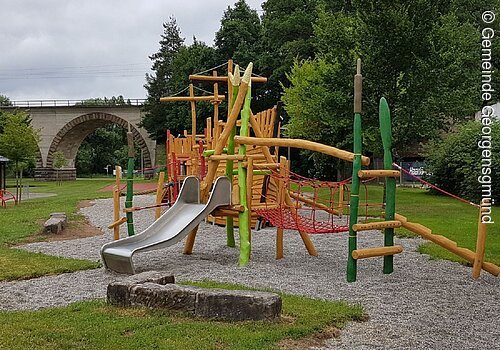 Ein Spieplatz mit großen Spielgeräten. Ein Klettergerüst mit Rutsche in bunten Farben. Im Hintergund grüne Bäume.