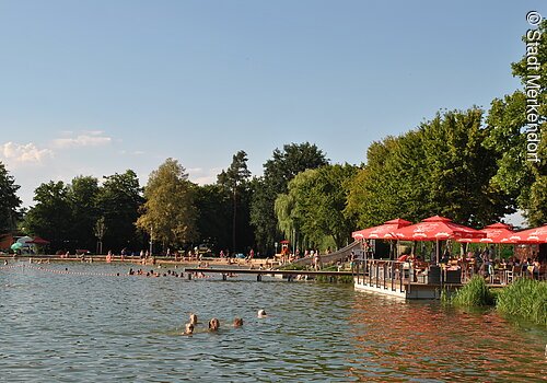 Ein Naturfreibad mit Steg. Im Wasser schwimmen Badegäste. Auf einer Holzterasse direkt am Wasser stehen rote Sonnenschirme.