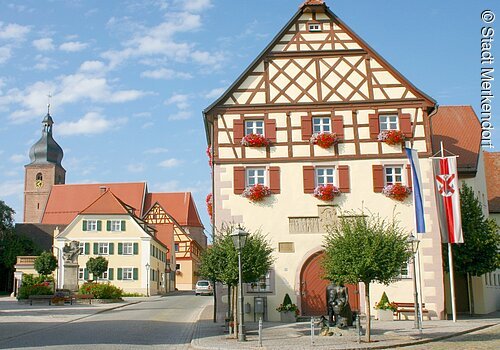 Im Fokus ein Rathaus im Fachwerkstil mit roten Balken und gelber Fassade. Vor dem Rathaus ein Krautbrunnen mit zwei Figuren und Krautsköpfen. Im Hintergund ragt der Kirchturm aus den Häusern der Altstadt.