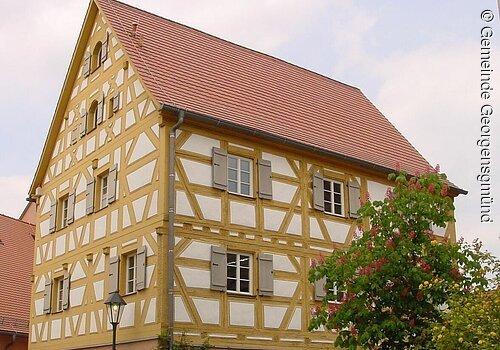 Ein Fachwerkhaus mit weißer Fassade und gelben Balken. Die Fensterläden sind grau. Da Dach aus roten Dachziegeln. Das Haus hat fünf Geschosse.