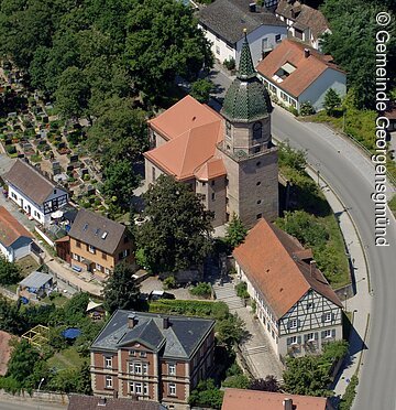 Luftbild eines Dorfes. In der Mitte eine Kirche mit grünem Turmdach. Drum herum Häuser und Bäume.