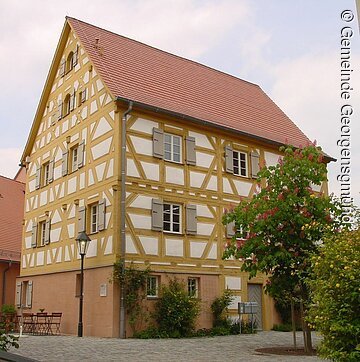 Fränkisches Fachwerkhaus Ein Fachwerkhaus mit weißer Fassade und gelben Balken. Die Fensterläden sind grau. Da Dach aus roten Dachziegeln. Das Haus hat fünf Geschosse.