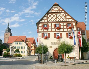 Im Fokus ein Rathaus im Fachwerkstil mit roten Balken und gelber Fassade. Vor dem Rathaus ein Krautbrunnen mit zwei Figuren und Krautsköpfen. Im Hintergund ragt der Kirchturm aus den Häusern der Altstadt.