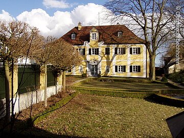 Ein Schloss im Barockstil mit weiß-gelber Fassade und grünen Fensterläden. Davor ein große Wiese.