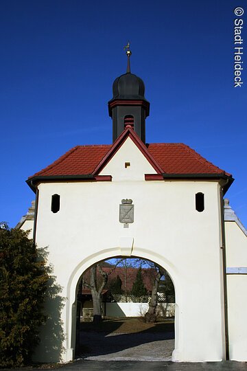 Ein Torbogen mit weißer Fassade und rotem Ziegeldach. Auf dem Dach ein kleines schwarzes Türmchen.