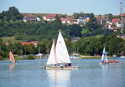 Ein weißes Segelschiff auf einem See. Daneben Windsurfer. Im Hintergund am Ufer Bäume. Etwas weiter hinten Wohnhäuser.