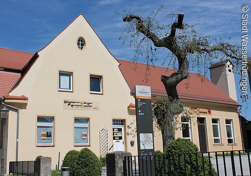 Ein Haus mit drei Ebenen und weißer Fassade. Davor ein Zaun. Über dem Eingang ein Schild mit der Aufschrift: Wassertrüdinger Figurentheater