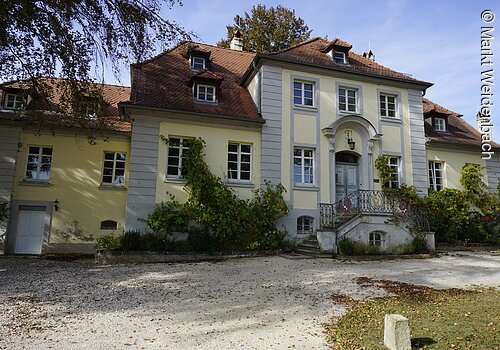 Ein barockes Gebäude mit gelber Fassade und rotem Ziegeldach. Das Haus hat mehere Unterteilungen. Am Eingang eine große Treppe die rechts und links von der Fassade hinauf zum Eingang führt.