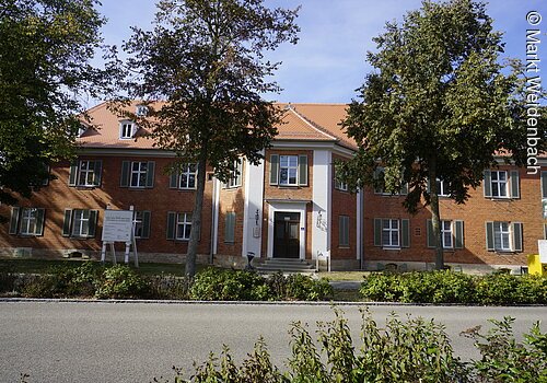 Frontansicht eines großen langen Gebäudes mit roter Ziegelsteinfassade. Das Schloss hat drei Ebenen. Der mittige Eingang ist baulich leicht hervorgehoben. An den Fenstern sind grüne Fensterläden befestigt.