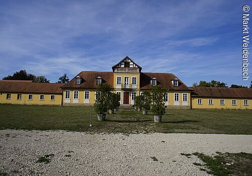 Ein langes barockes Gebäude. Gelbe Fassade mit rotem Ziegeldach. Davor eine große Wiese.