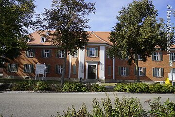 Frontansicht eines großen langen Gebäudes mit roter Ziegelsteinfassade. Das Schloss hat drei Ebenen. Der mittige Eingang ist baulich leicht hervorgehoben. An den Fenstern sind grüne Fensterläden befestigt.