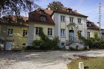 Ein barockes Gebäude mit gelber Fassade und rotem Ziegeldach. Das Haus hat mehere Unterteilungen. Am Eingang eine große Treppe die rechts und links von der Fassade hinauf zum Eingang führt.