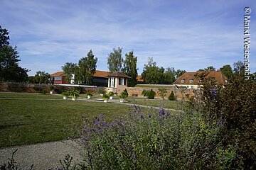 Ein Garten mit großer grüner Wiese. Am Rand des Gartens eine Mauer aus roten Ziegeln mit kleinem Häuschen.