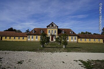Ein langes barockes Gebäude. Gelbe Fassade mit rotem Ziegeldach. Davor eine große Wiese.