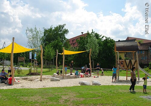 Ein Spielplatz auf Schotterfläche umgeben mit grüner Wiese. Zwei kleine gelbe Sonnensegel. Rechts daneben eine Rutsche. Kinder und Eltern spielen auf dem Spielplatz. Im Hintergund Büsche und Bäume.