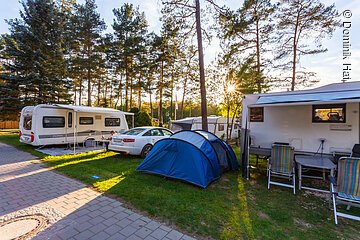 Ein Campingplatz bei Sonnenuntergang. Auf einer grünen Wiese stehen ein Wohnwagen, daneben ein weißes Auto, dann ein blaues Zelt und ganz rechts ein weiterer Wohnwagen.