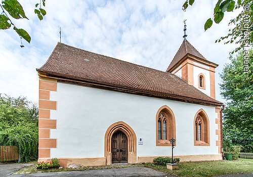 Außenansicht einer Kapelle mit kleinem Turm. Weiße Fassade mit orangenen Akzenten und rotem Ziegeldach.