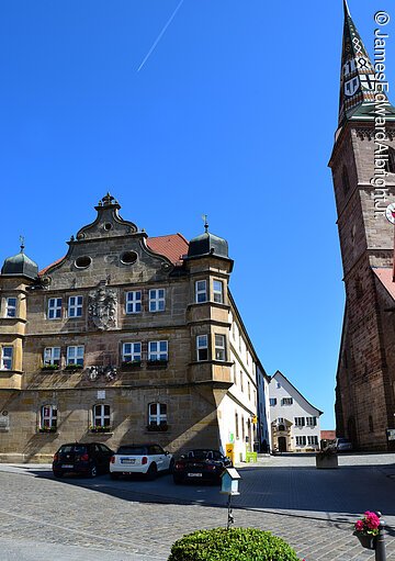 Blick auf ein Deutschordenschloss. Ein Haus mit vier Stöcken und Steinfassade. Das Schloss steht in einer gepflasterten Altstadt. Rechts ein Kirchturm mit buntem Ziegeldach.