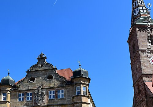 Blick auf ein Deutschordenschloss. Ein Haus mit vier Stöcken und Steinfassade. Das Schloss steht in einer gepflasterten Altstadt. Rechts ein Kirchturm mit buntem Ziegeldach.