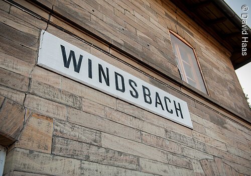 Nahaufnahme einer Hausfassade. An der Steinfassade hängt ein weißes Schild. Darauf in schwarzen Blockbuchstaben das Wort Windsbach.