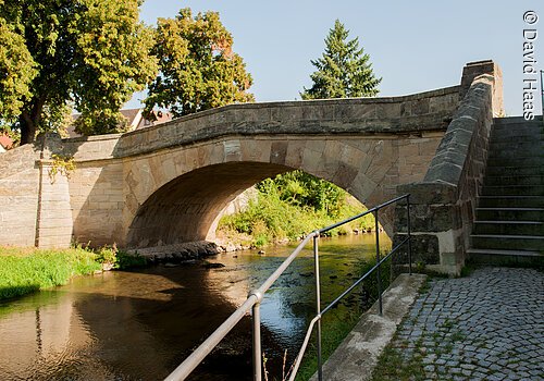 Eine Steinbrücke führt über einen Fluss. Im Hintergrund Bäume.