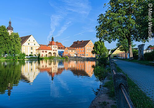 Teil eines Weihers. Im Wasser spiegelns ich die Häuserfassaden einer Stadt. Die Fassaden werden von der Sonne angestrahlt. Rechts verläuft ein Fußgängerweg entlang des Weihers.