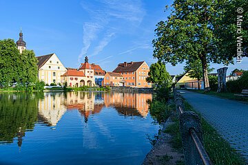 Teil eines Weihers. Im Wasser spiegelns ich die Häuserfassaden einer Stadt. Die Fassaden werden von der Sonne angestrahlt. Rechts verläuft ein Fußgängerweg entlang des Weihers.