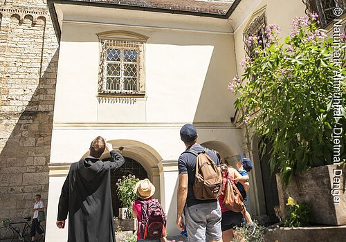 Eine Familie steht mit einem Mönch vor dem Eingang zum Kloster Plankstetten. Der Mönch erklärt der Familie etwas von der Geschichte des Gebäudes. Er hat seinen Arm gehoben und zeigt auf etwas.