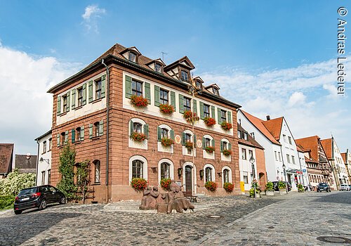 Ein großes Haus mit vier Ebenen. Ein Haus mit Steinfassade. An den Fenstern grüne Fensterläden. Daran Blumenkästen mit roten Blumen. Das Haus hat ein Walmdach. Das Haus liegt an einer gepflasterten Straße.