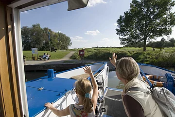 Eine Frau und ein junges Mädchen winken vom Schiff den Besuchern am grünen Uferrand.