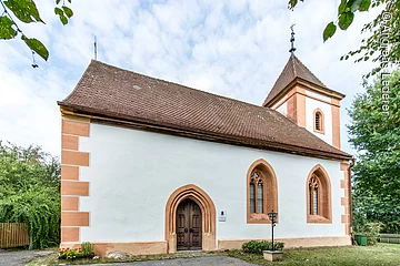 Außenansicht einer Kapelle mit kleinem Turm. Weiße Fassade mit orangenen Akzenten und rotem Ziegeldach.