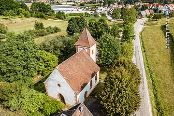 Luftaufnahme eienr Kappekke mit kleinem Turm. Kapelle mit weißer Fassade und rotem Ziegeldach. Die Kapelle ist umgeben von grünen Bäumen und Sträuchern.