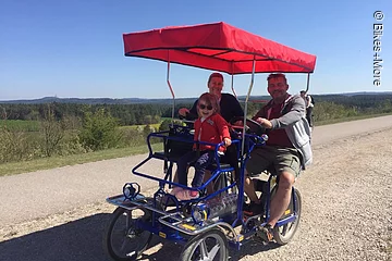 Eine Ehepaar mit Kind fährt auf einer Rikscha auf einem Schotterweg durch eine grüne Landschaft. Die Familie lächelt in die Kamera.