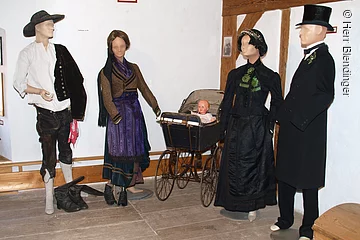 Vier Schaufensterpuppen stehen in einem Raum mit weißer Wand und braunem Fachwerk. Die Puppen tragen fränkische Trachten. In der Mitte ein alter Kinderwagen.